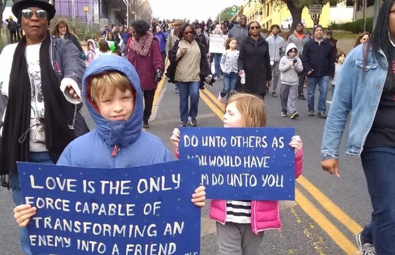 depicts many diverse people marching for change. Two small boys hold signs that say, "Love is the only force capable of transforming an enemy into a friend" and "do unto others as you would have them do unto you.