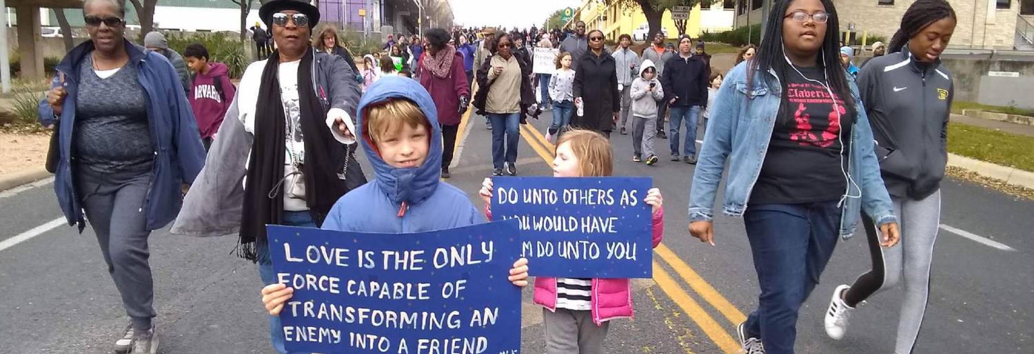 depicts many diverse people marching for change. Two small boys hold signs that say, "Love is the only force capable of transforming an enemy into a friend" and "do unto others as you would have them do unto you.